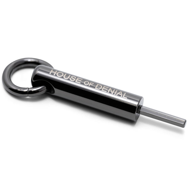 A black cylinder shaped pendant with a loop through it, a hexagon key end and House of Denial printed along the body.