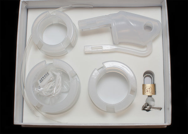 Clear HoD100 silicone chastity kit with all of the individual parts laid out inside the foam lining of the storage box. It shows the cage, 3 back rings, 2 plastic locking strips, the cage and a brass padlock with 3 keys.
