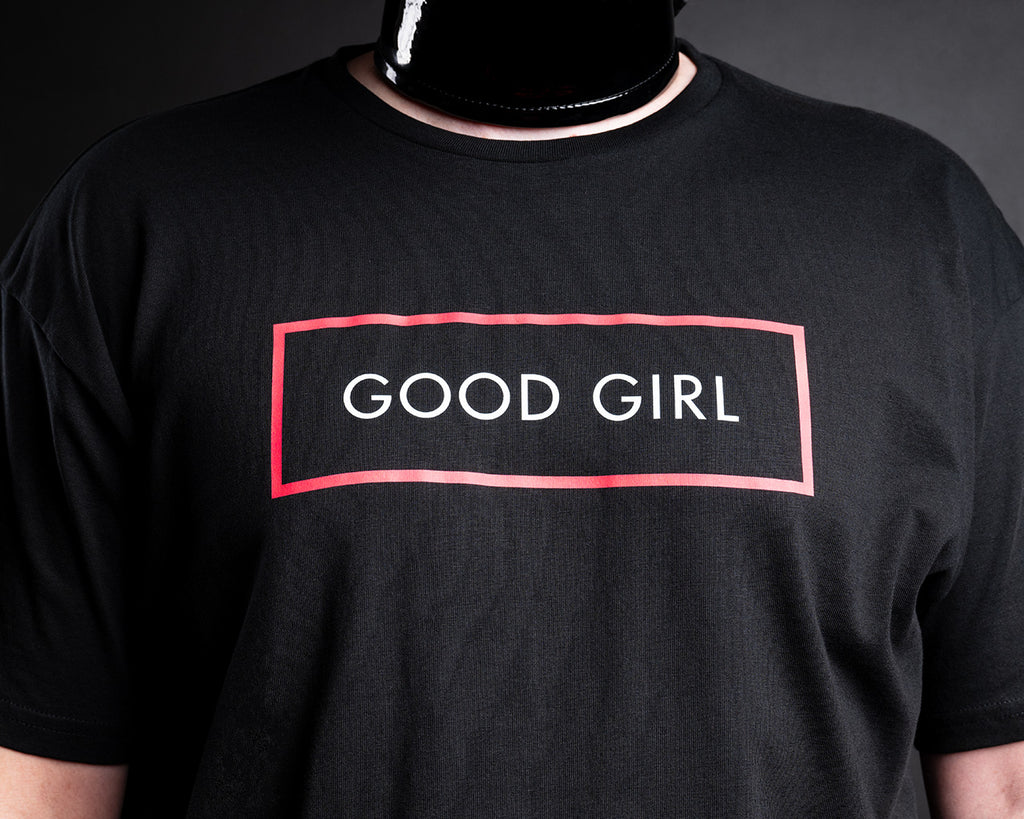 A close-up of slave D wearing a black short-sleeve t-shirt with a red rectangular outline and the words 'GOOD GIRL' printed in white across the chest.