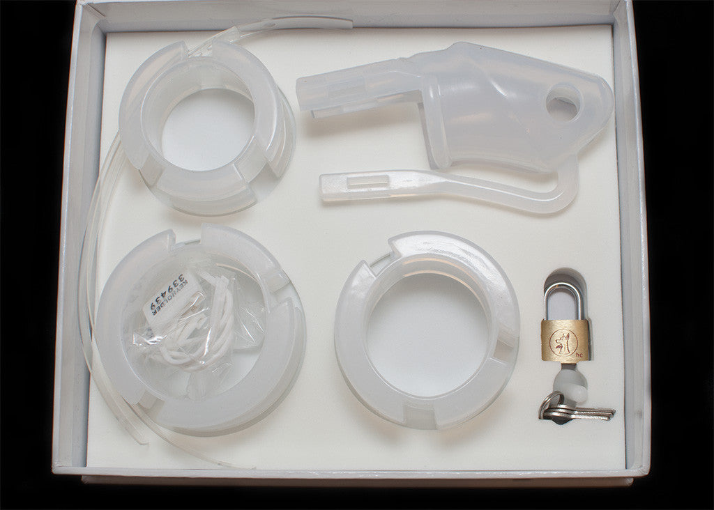 Clear HoD100 silicone chastity kit with all of the individual parts laid out inside the foam lining of the storage box. It shows the cage, 3 back rings, 2 plastic locking strips, the cage and a brass padlock with 3 keys.